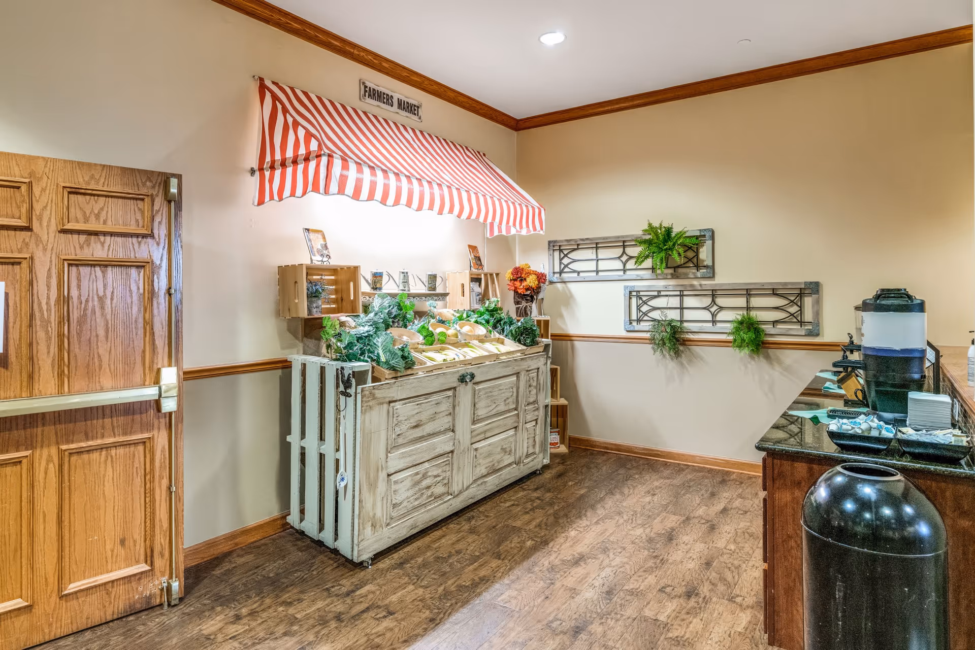 Indoor corner in a senior living facility featuring a rustic farmers market display with produce under a red-and-white awning and a nearby coffee station.