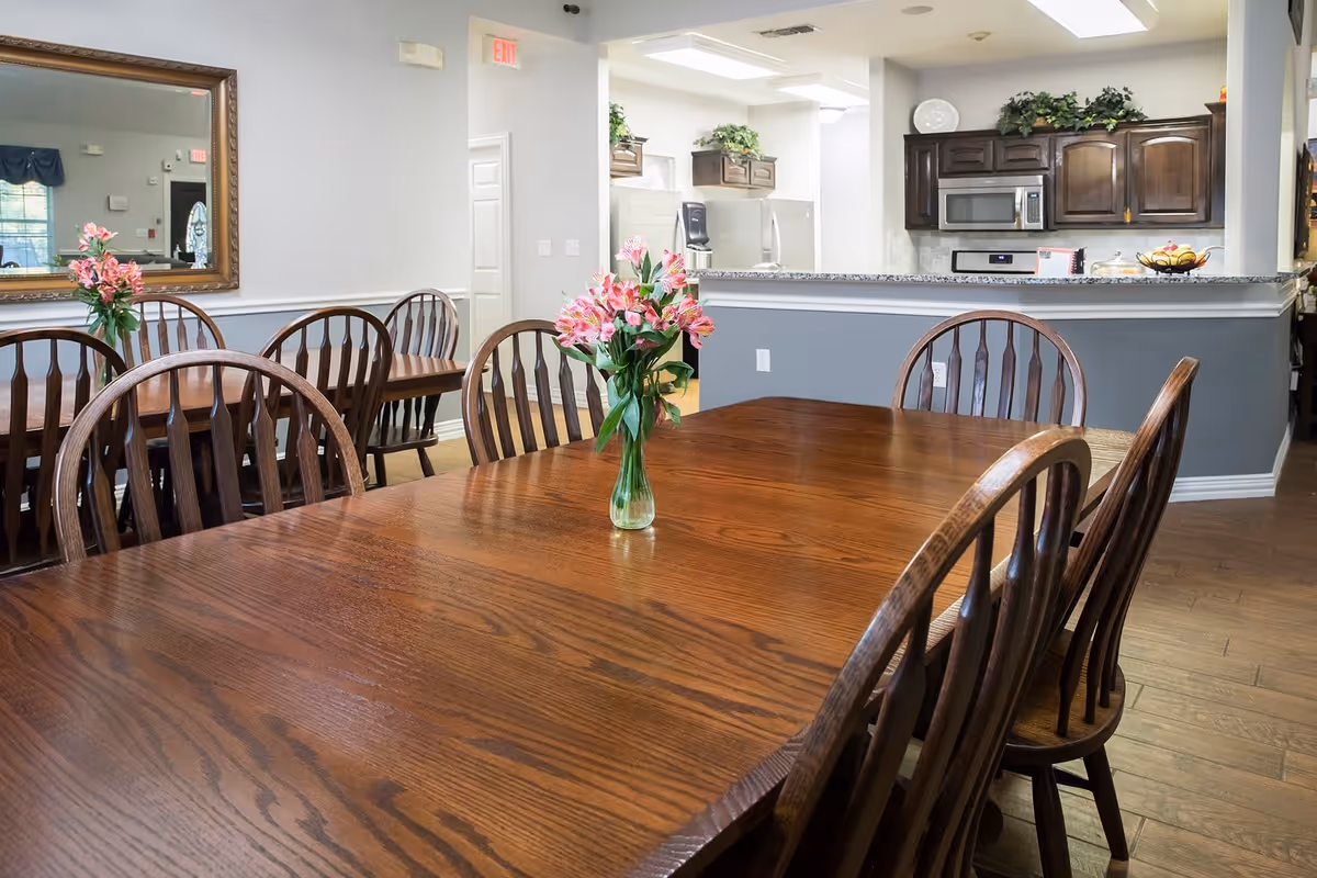 A dining area with wooden tables and chairs, each table decorated with a vase of pink flowers. In the background, there is a kitchen with dark wooden cabinets, a microwave, and a refrigerator. The walls are painted light gray with white trim, and there is a large mirror on one wall reflecting part of the room.