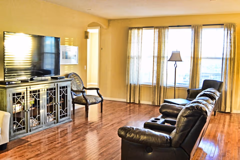 A bright living room with wooden flooring, featuring a large flat-screen TV on a decorative cabinet, a patterned armchair, a leather reclining sofa, a floor lamp, and large windows with sheer curtains allowing natural light to fill the space.
