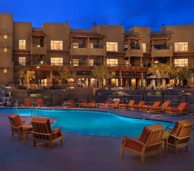 Evening view of an outdoor swimming pool area with lounge chairs and wooden seating. Behind the pool is a multi-story building with balconies and warm interior lighting, surrounded by trees and landscaping.