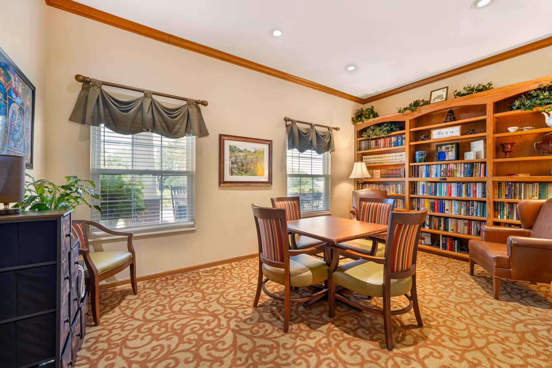 Cozy communal library/lounge with a central table and chairs, bookshelves along the wall, armchairs, windows, and warm decor.