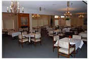 A spacious dining room with multiple tables covered with white tablecloths, each surrounded by wooden chairs with cushioned seats. The room features chandeliers hanging from the ceiling, framed artwork on the walls, and large windows with blinds.