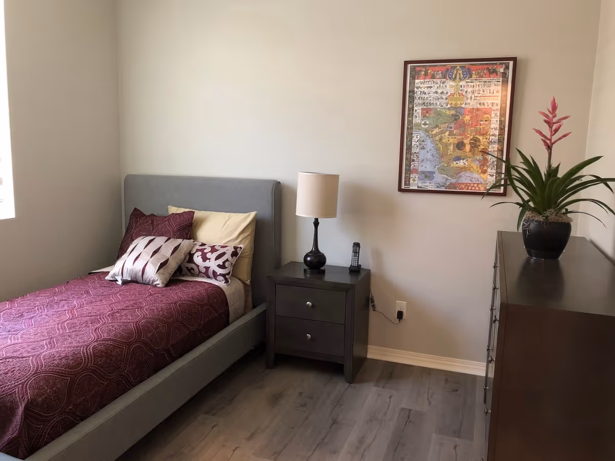 A neatly made single bed with a gray upholstered headboard, covered with a purple patterned bedspread and multiple pillows. Next to the bed is a dark wooden nightstand with a lamp and a cordless phone. On the right side, there is a dark wooden dresser with a potted plant on top. A framed colorful artwork hangs on the wall above the nightstand. The room has light-colored walls and wood flooring.