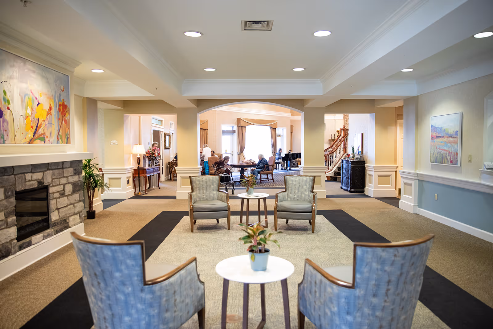 A bright and spacious common area in a senior living facility with comfortable seating arranged around small tables. The room features a stone fireplace on the left, colorful artwork on the walls, and a carpeted floor. In the background, several elderly people are seated at tables, engaging in activities, with large windows letting in natural light.