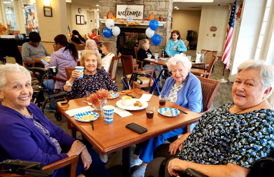 Four elderly women sitting around a table in a dining/common area of a senior living facility, smiling and holding drinks.