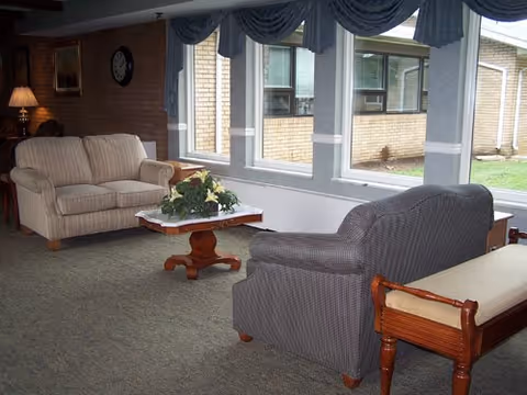A cozy living room area with two upholstered sofas facing each other across a wooden coffee table with a floral centerpiece. Large windows with blue valances let in natural light and show an exterior view of a brick building and grass. A wooden bench with a cushioned seat is positioned near one sofa, and a lamp and clock are visible on the far wall.