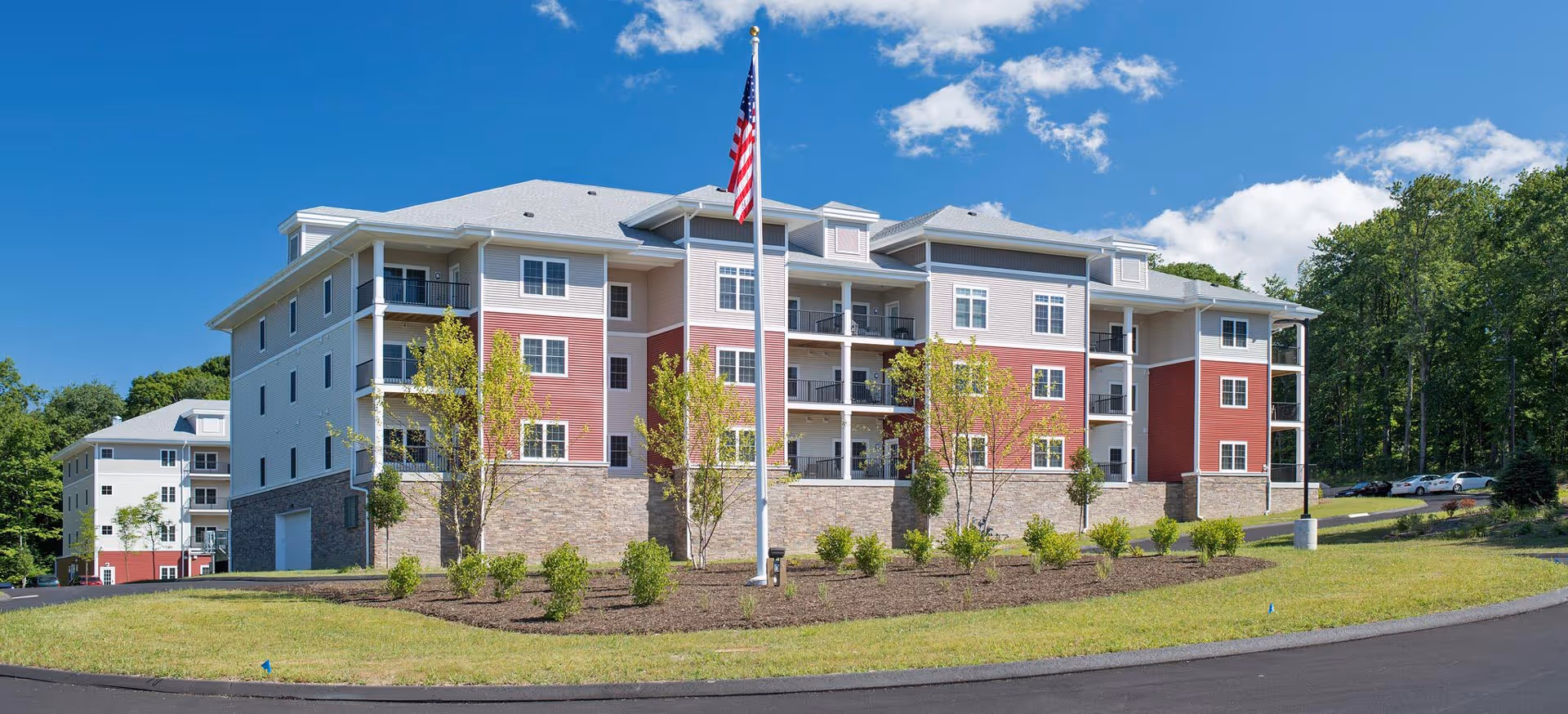 Exterior view of a multi-story senior living facility building with red and beige siding, balconies, and a stone foundation. There is a flagpole with an American flag in front, surrounded by landscaped bushes and trees under a blue sky with some clouds.