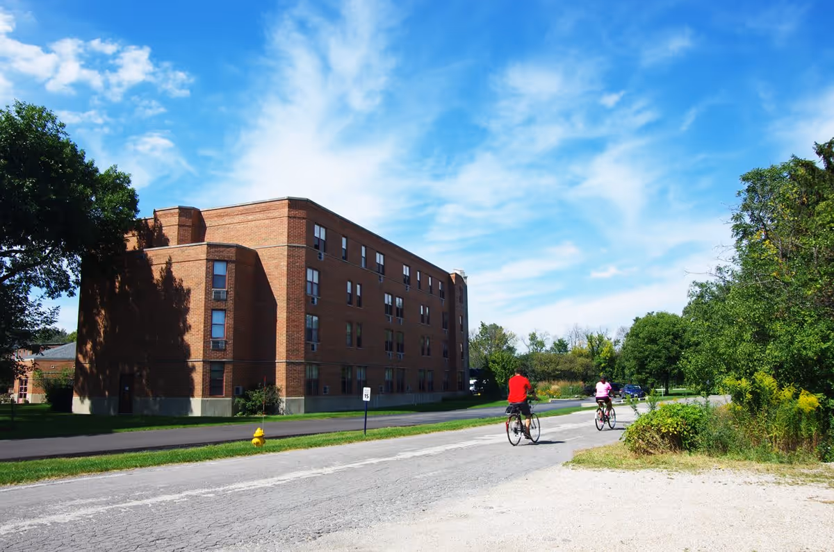 A large brick multi-story building under a blue sky with scattered clouds. Two people are riding bicycles on a paved road in front of the building. There are trees and greenery on both sides of the road.