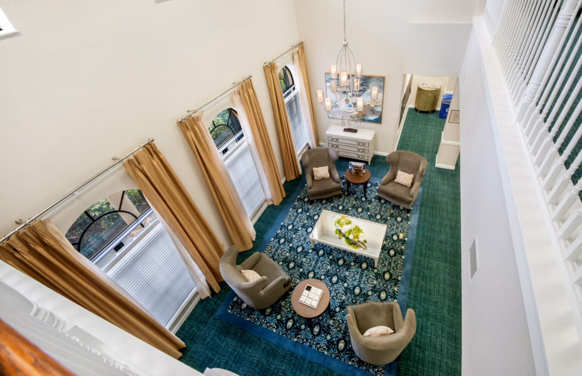 A bright and spacious living room area viewed from above, featuring four gray armchairs arranged around a patterned blue area rug with a white coffee table in the center. The room has tall windows with beige curtains, a modern chandelier, a white sideboard with decorative items, and white walls with a green carpeted floor.