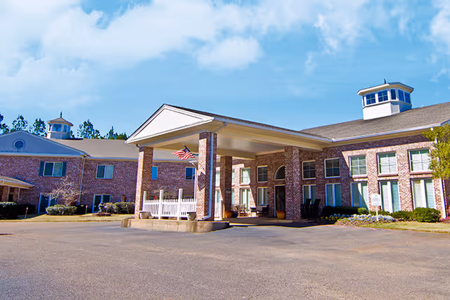 Brick senior living facility with a covered entrance porte-cochere, American flag, and two-story wings under a blue sky.