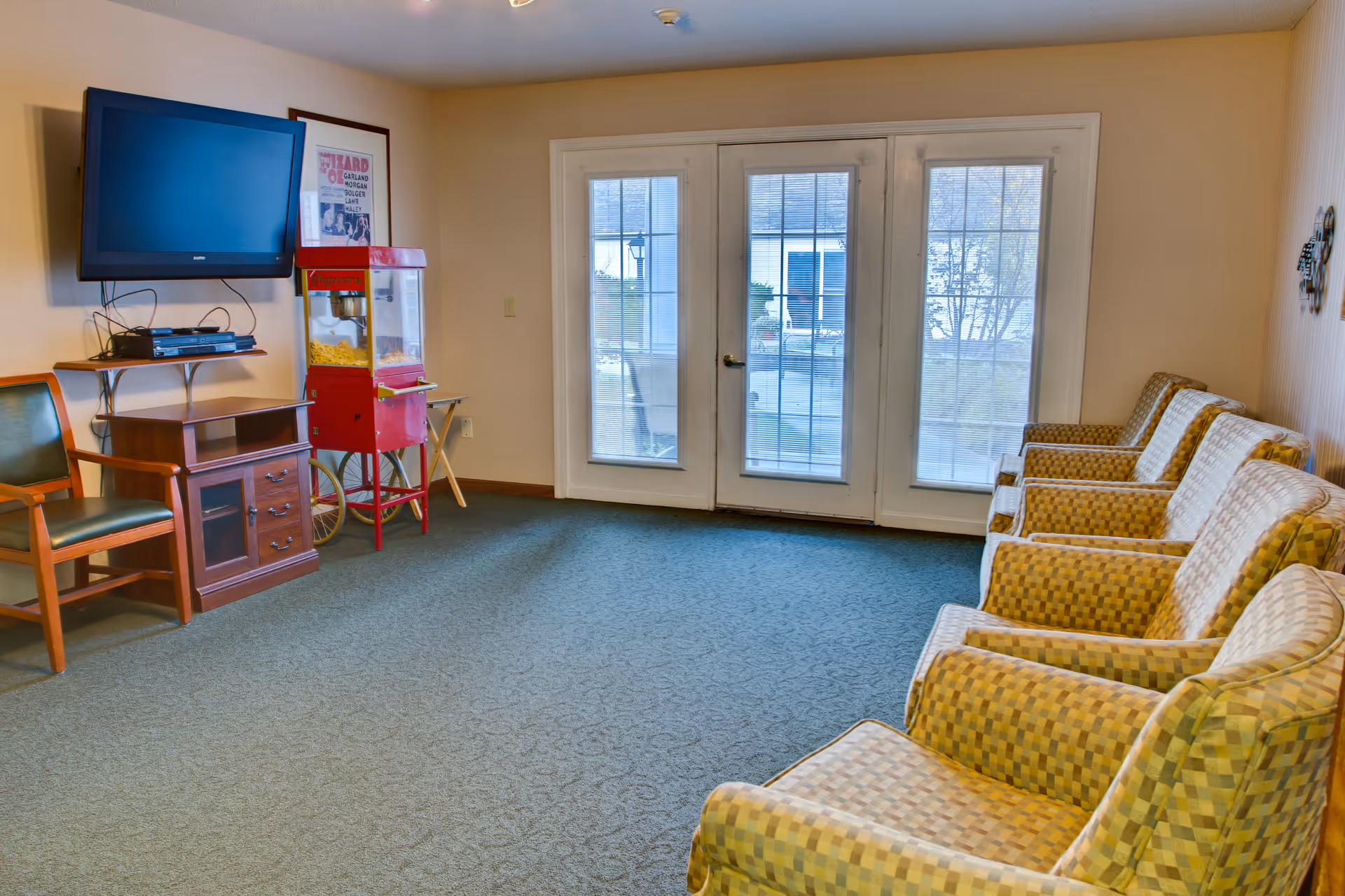 A common lounge area with a row of patterned armchairs on the right side, a wall-mounted flat screen TV on the left, a wooden cabinet below the TV, and a red popcorn machine next to the cabinet. There are glass double doors with blinds leading outside at the back of the room.