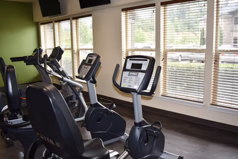 Interior view of a fitness room with several exercise bikes positioned in front of large windows with blinds. The room has light-colored walls and a green accent wall, with a television mounted near the ceiling.