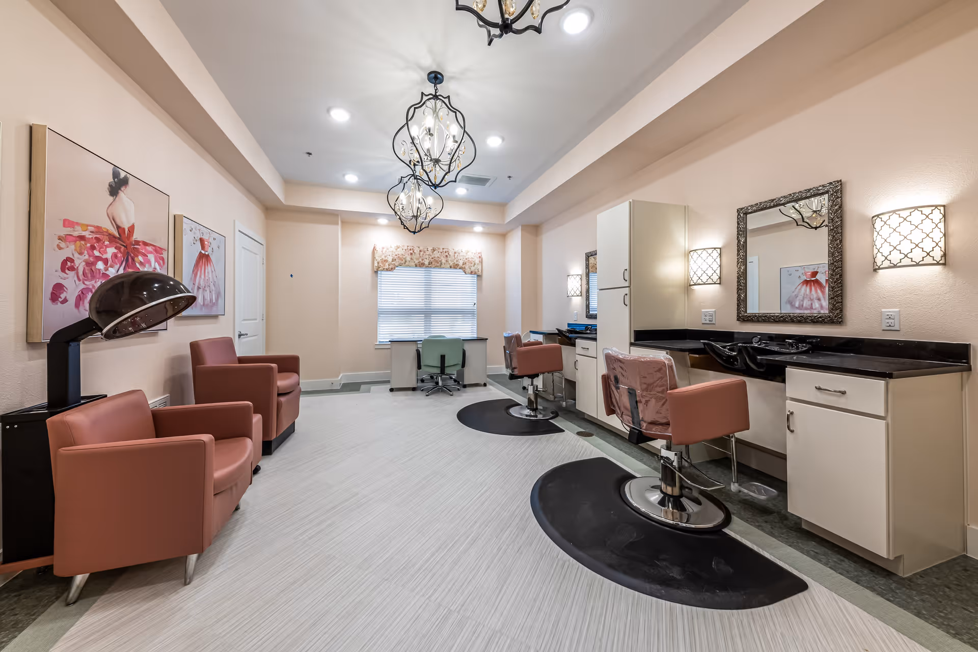 A bright and clean salon area in an assisted living facility featuring two salon chairs with mirrors and sinks, two comfortable armchairs, a hair dryer, and a desk with a chair near a window with blinds and floral valance. The walls are painted light peach, and decorative paintings and light fixtures add a cozy ambiance.