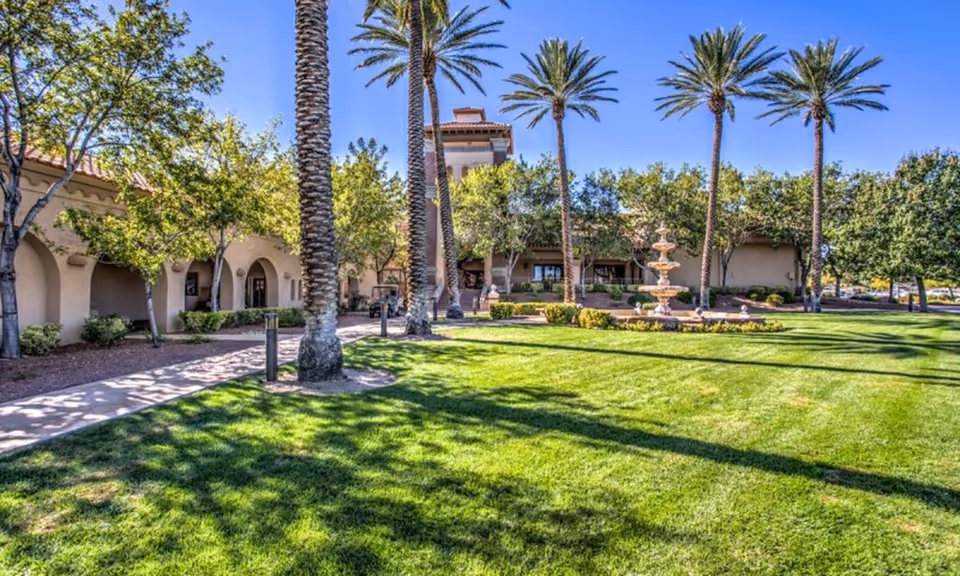 Sunny courtyard with palm trees, a green lawn and a fountain in front of a Mediterranean-style clubhouse.