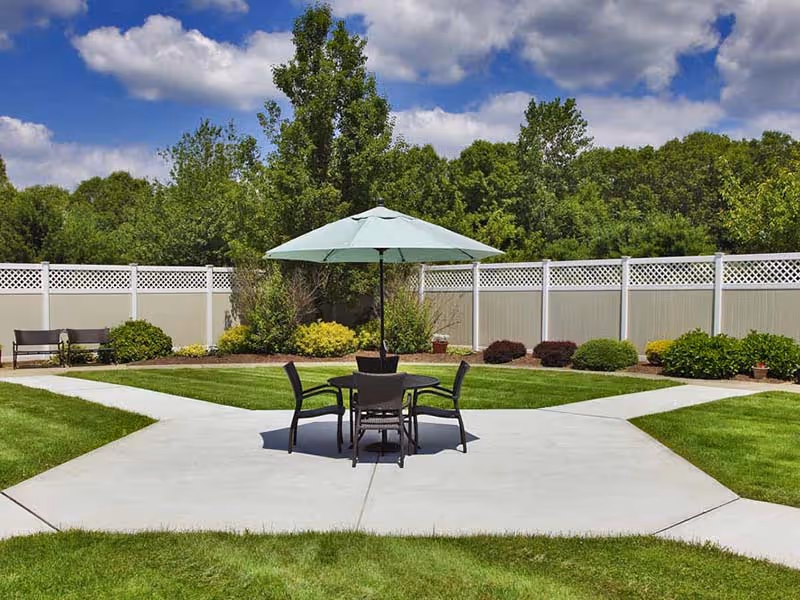 Outdoor patio area with a round table, four chairs, and a large umbrella in the center, surrounded by green grass, bushes, and trees with a white fence enclosing the space under a partly cloudy blue sky.