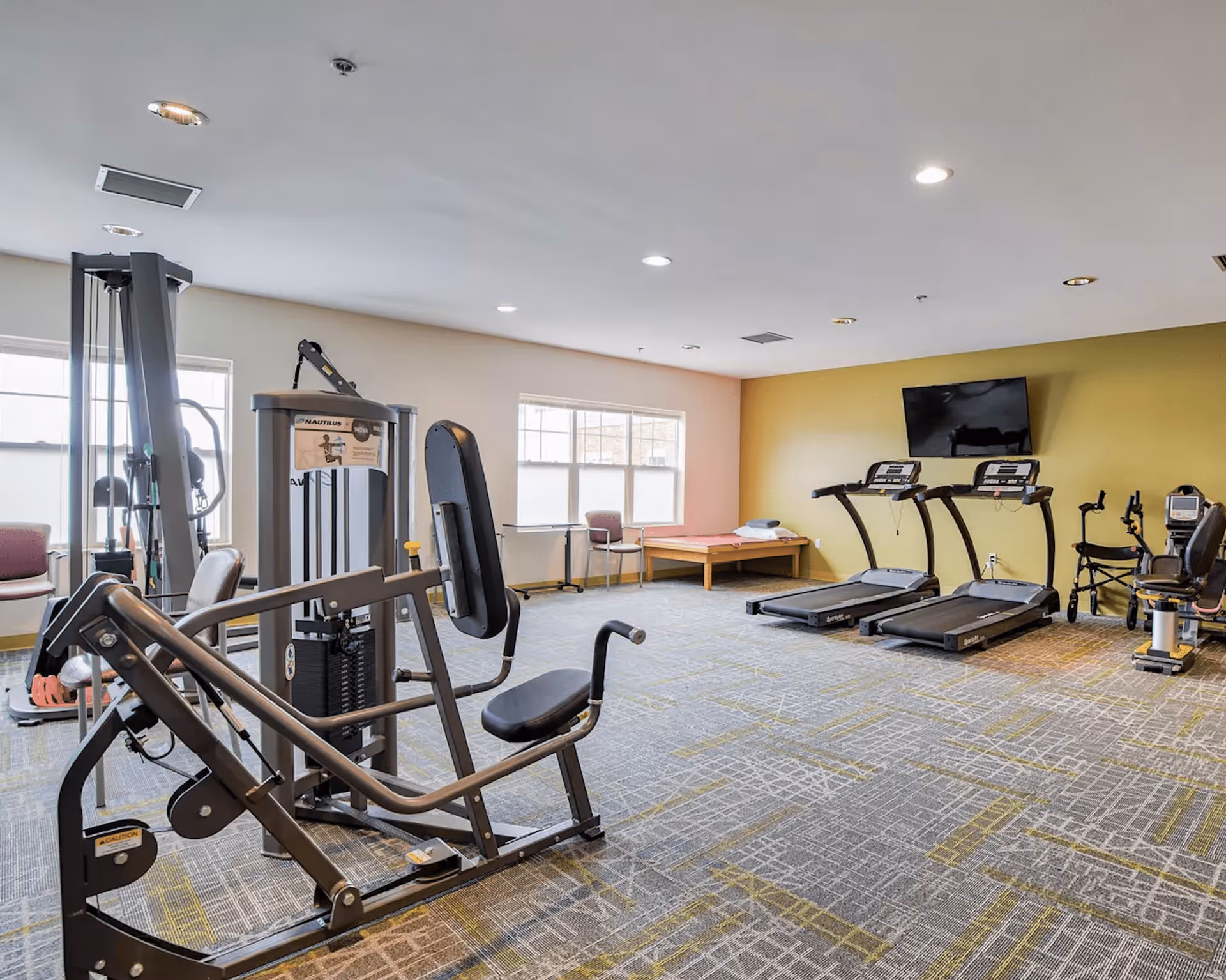 A well-lit fitness room with exercise equipment including a weight machine, two treadmills, and a recumbent bike. The room has large windows, a yellow accent wall with a mounted flat-screen TV, and a padded bench with folded towels.