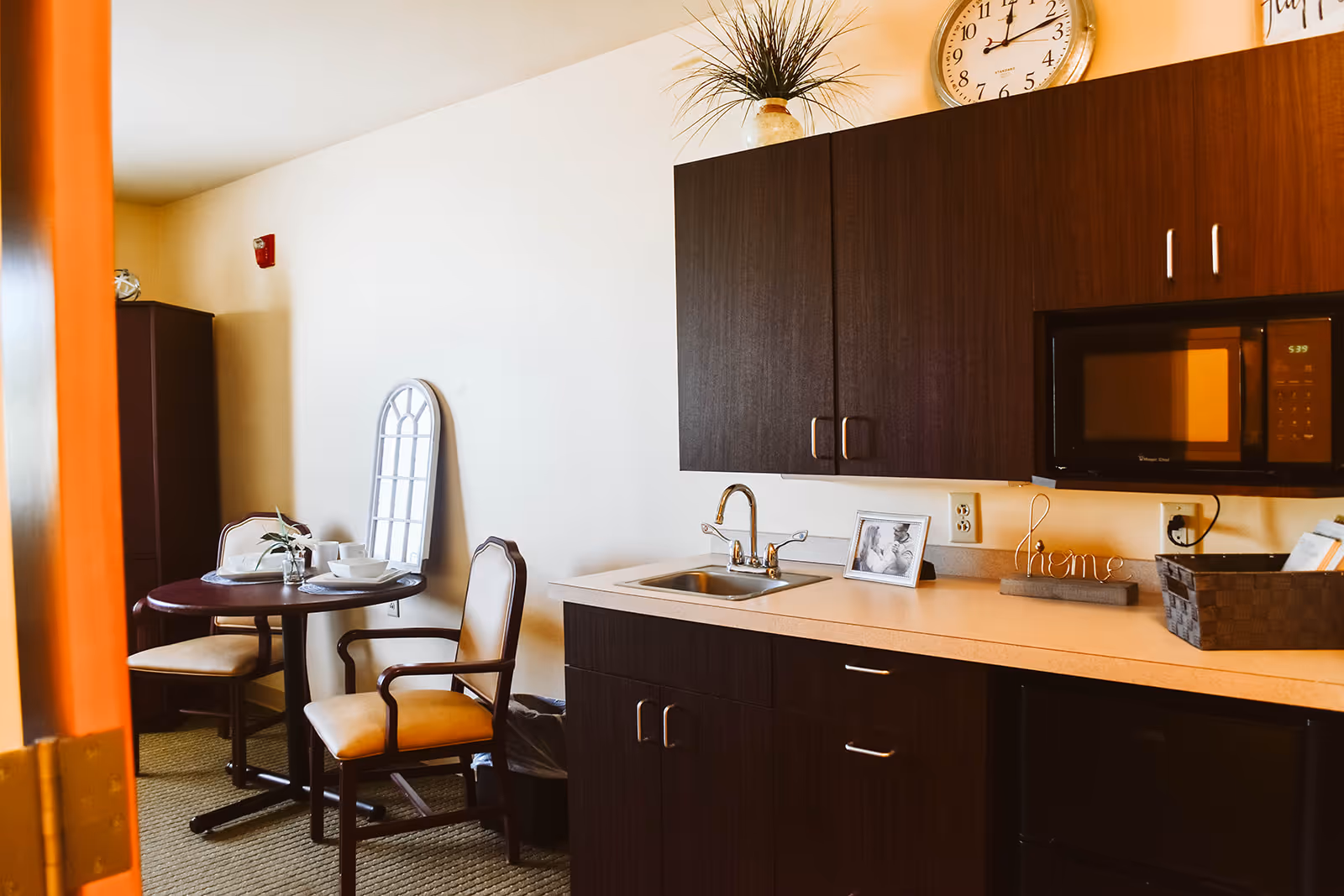 A small kitchenette area with dark wood cabinets, a sink, a microwave, and a mini refrigerator. To the left, there is a round dining table with two chairs, set with white dishes and a small plant. A decorative mirror leans against the wall behind the table, and a clock and plant are on top of the cabinets.