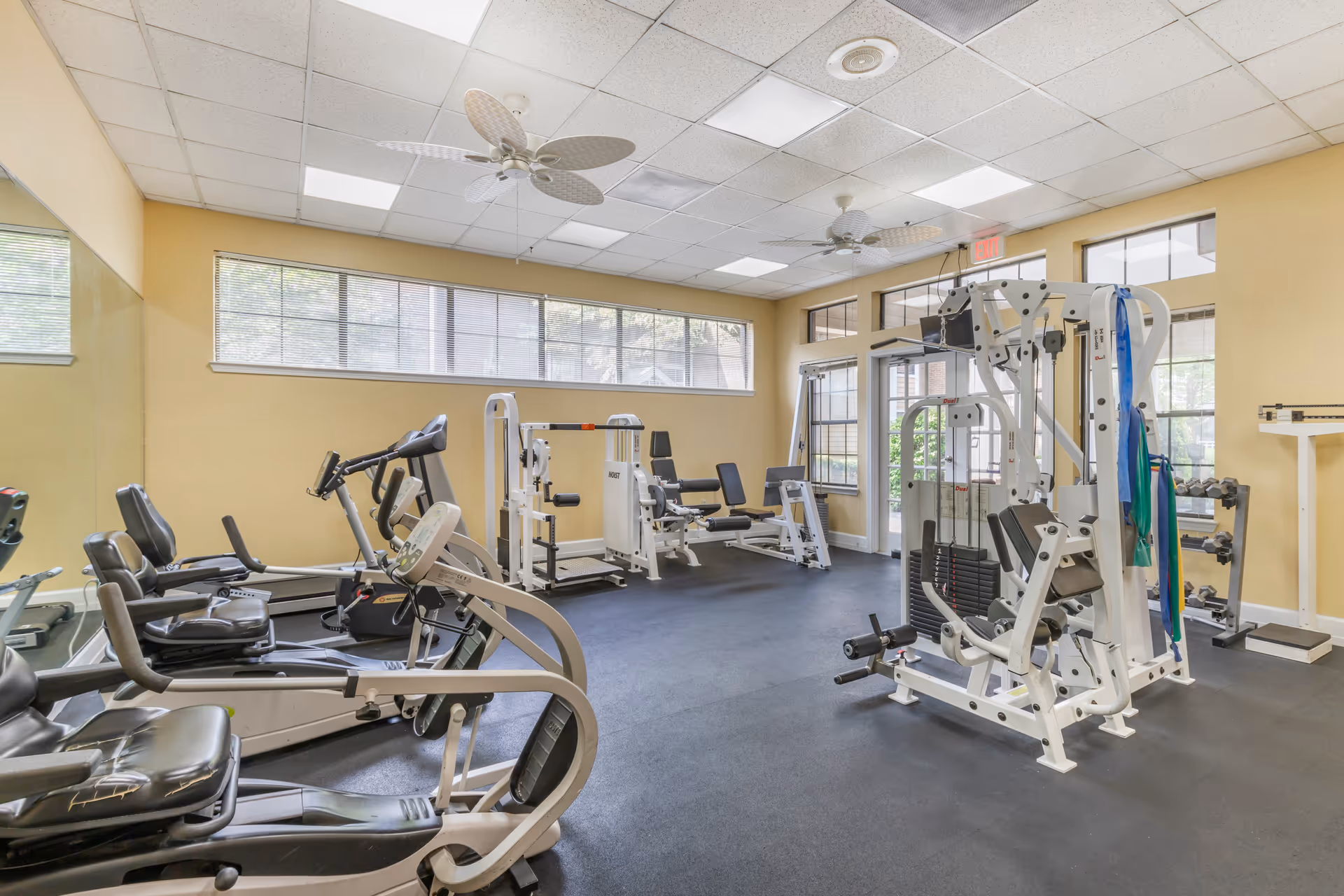 A well-lit fitness room with various exercise equipment including recumbent bikes, weight machines, and resistance bands. The room has large windows, ceiling fans, and a mirror on one wall.