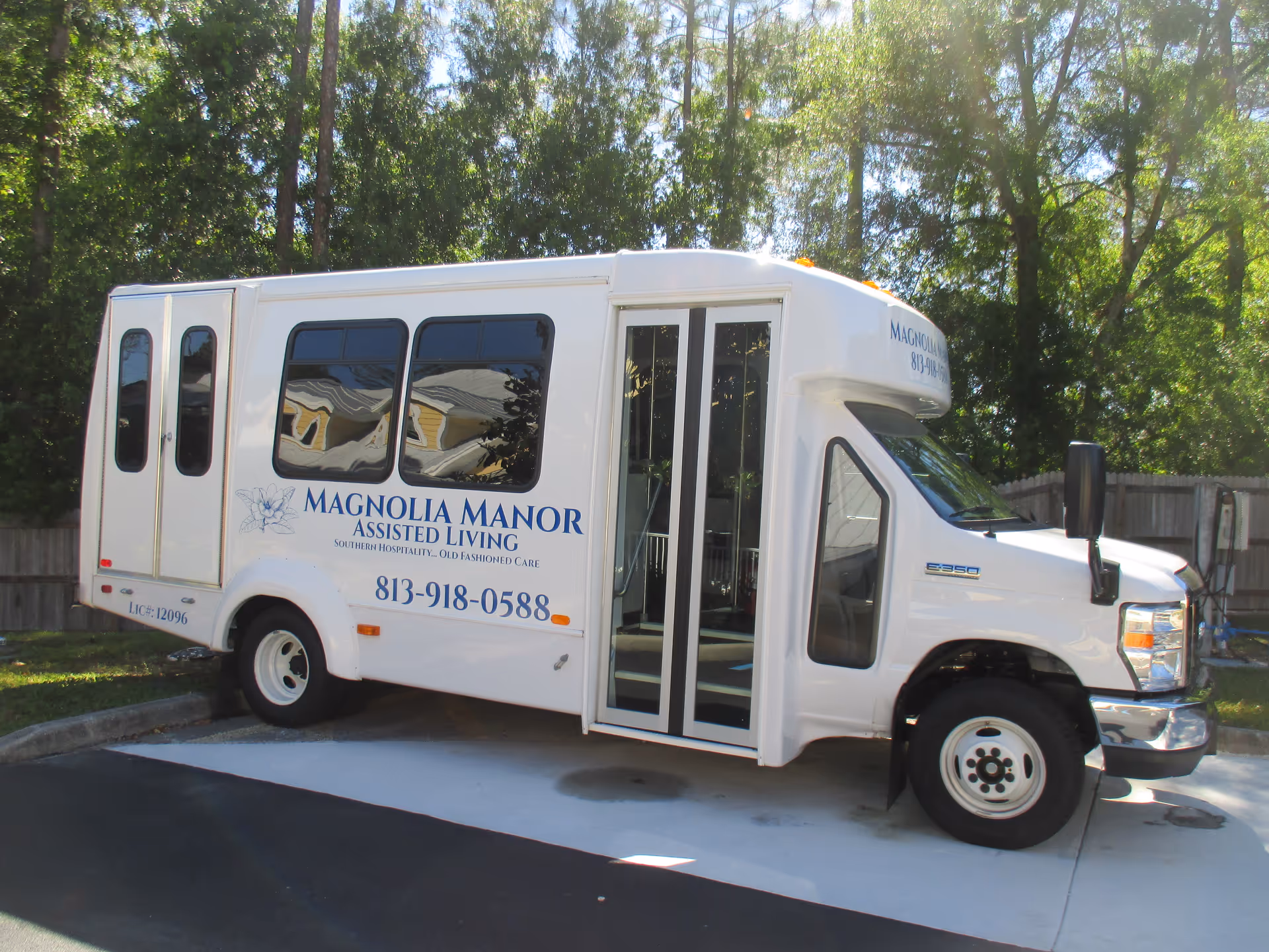 A white Magnolia Manor Assisted Living shuttle bus parked outdoors beside trees and a fence.