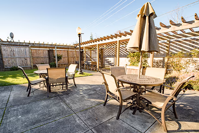Sunny outdoor courtyard with round tables and chairs, umbrellas, a pergola, and a wooden fence.