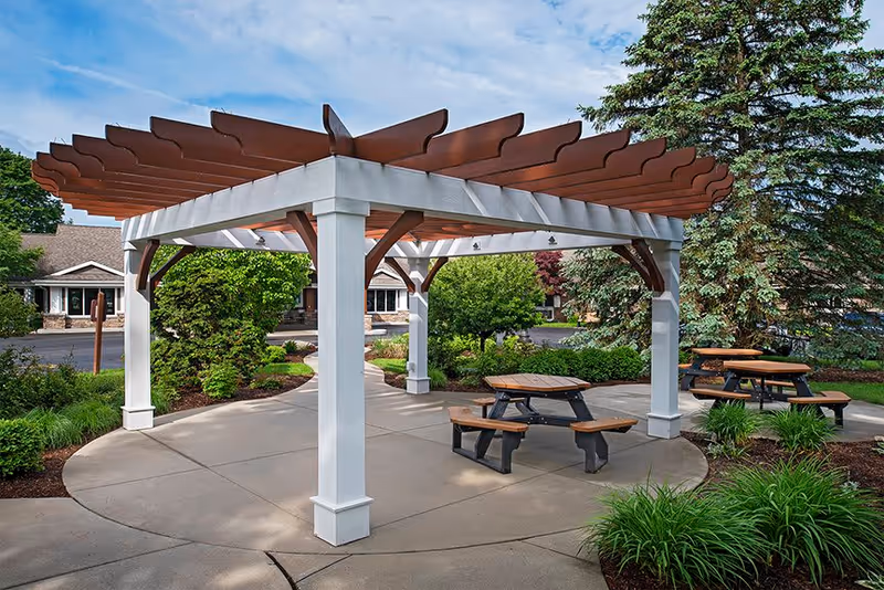 Outdoor seating area with wooden picnic tables and benches under a white pergola with brown beams, surrounded by green shrubs, trees, and a paved walkway in a garden setting at American House Kentwood.
