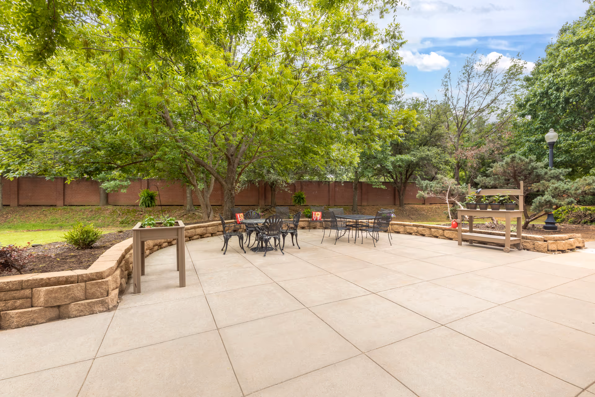 Sunny outdoor patio with metal tables and chairs, raised stone planters, and large trees in the background.