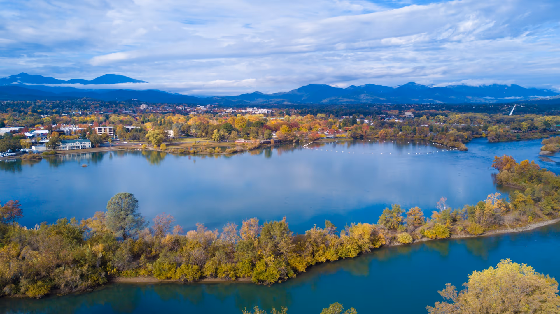 Aerial view of a large lake surrounded by trees with autumn foliage, a town in the background, and mountains under a partly cloudy sky.