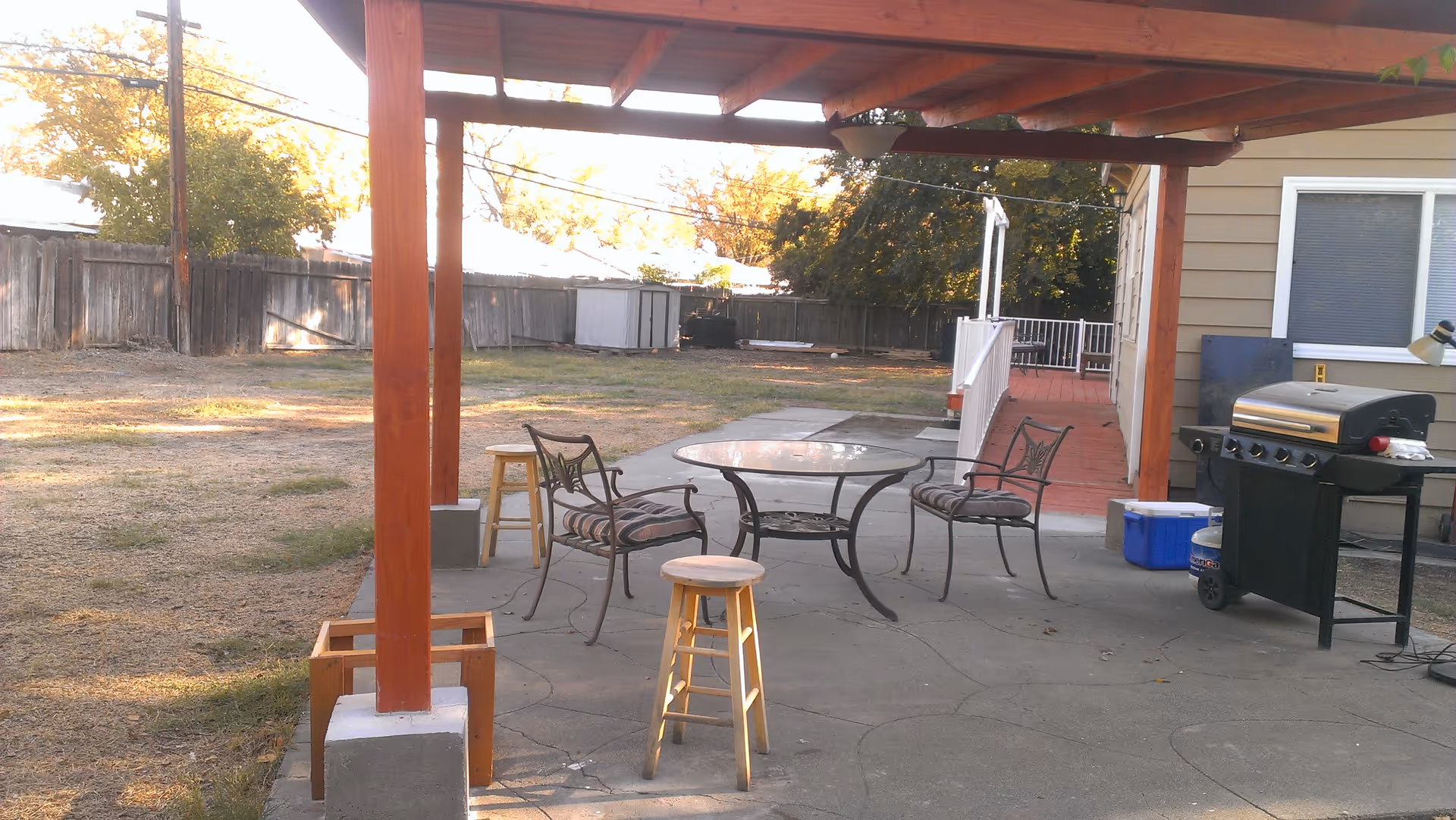 Backyard patio under a wooden pergola with a glass table, metal chairs and stools, a grill, and a grassy yard beyond.