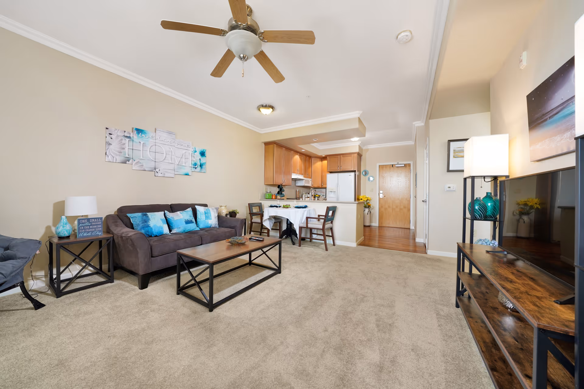 A bright and cozy living room area with beige walls and carpet. The room features a brown sofa with blue and white cushions, a wooden coffee table, and a side table with a lamp and decorative items. A ceiling fan with wooden blades is mounted on the ceiling. In the background, there is a small dining table with two chairs and a kitchen area with wooden cabinets, a refrigerator, and a stove. A flat-screen TV is placed on a wooden TV stand with shelves, and a floor lamp with a white shade stands beside it. The entrance door is visible at the far end of the room.