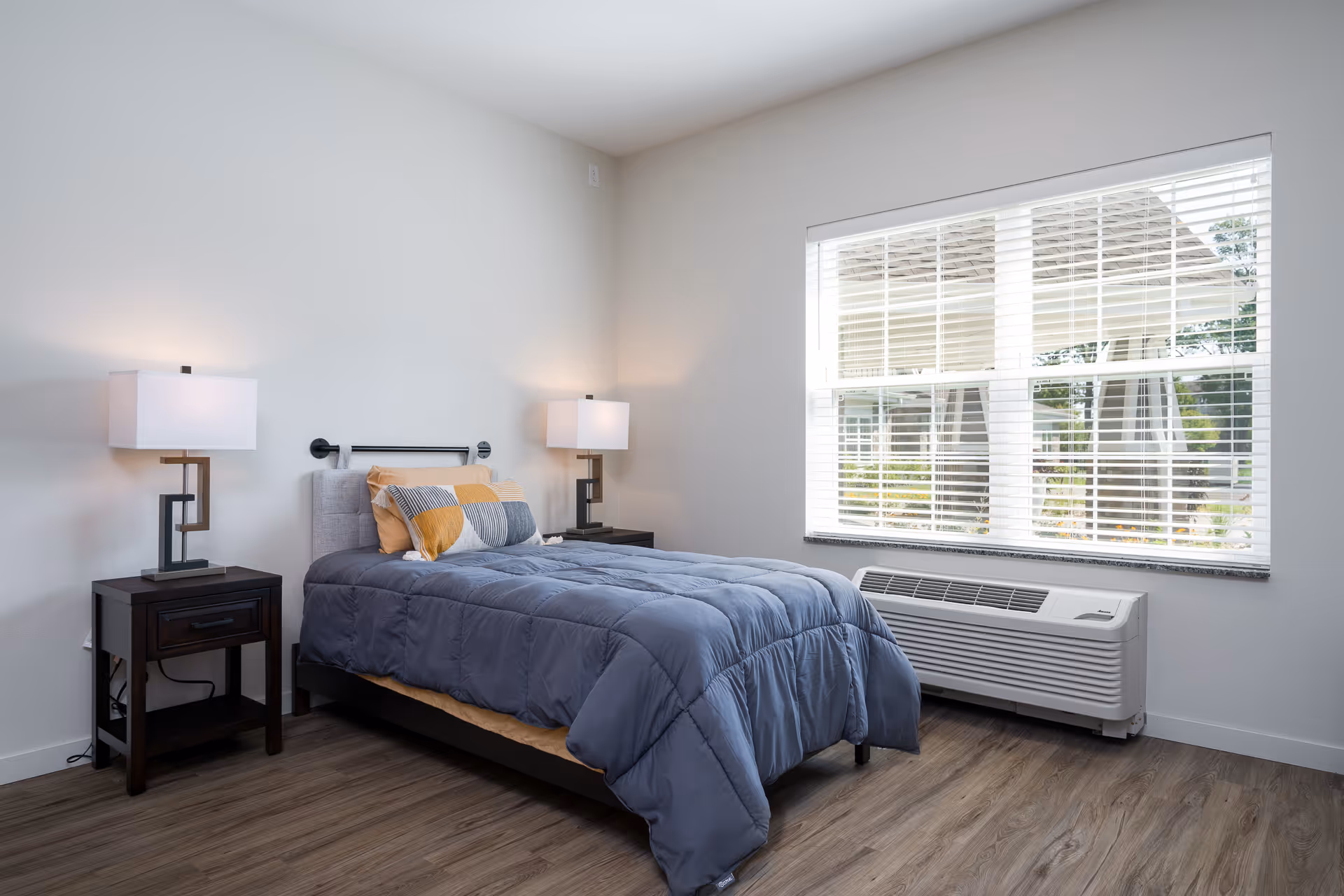 A neatly made single bed with a blue comforter and decorative pillows in a bright bedroom. The room features two modern bedside tables with matching lamps, a large window with white blinds, and a wall-mounted air conditioning unit below the window. The floor is wood, and the walls are painted white.