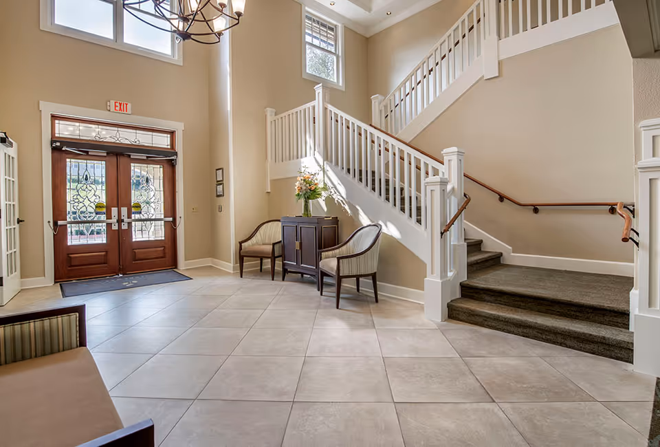 Bright foyer with double wooden entrance doors, tiled floor, a staircase with white railing, and a small seating area.
