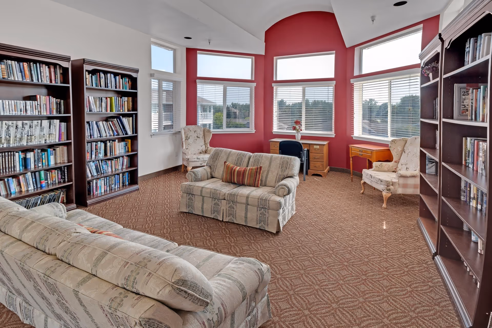 A bright and cozy library or reading room with large windows letting in natural light. The room features two patterned sofas with a striped pillow, two floral armchairs, wooden bookshelves filled with books, and two wooden desks with chairs positioned near the windows. The walls are painted red and white, and the floor is carpeted with a geometric pattern.