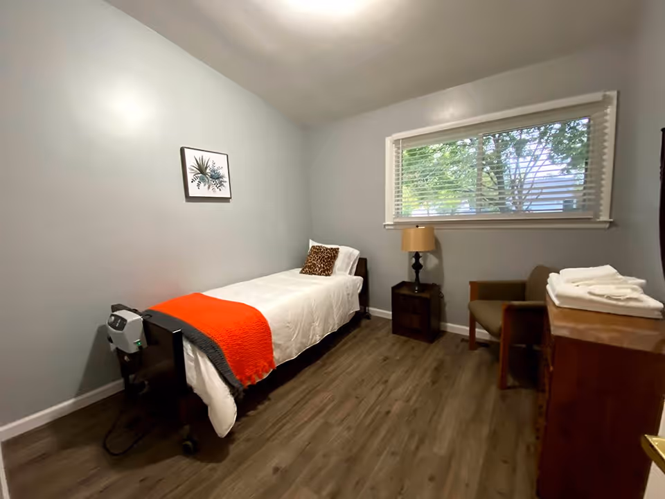 A small, simple bedroom with a single bed covered in white bedding and an orange throw blanket. There is a leopard print pillow on the bed. Next to the bed is a small wooden nightstand with a lamp. A window with blinds shows green trees outside. There is a wooden dresser with folded white towels on top and a brown armchair beside it. The walls are painted light gray and the floor is wood.