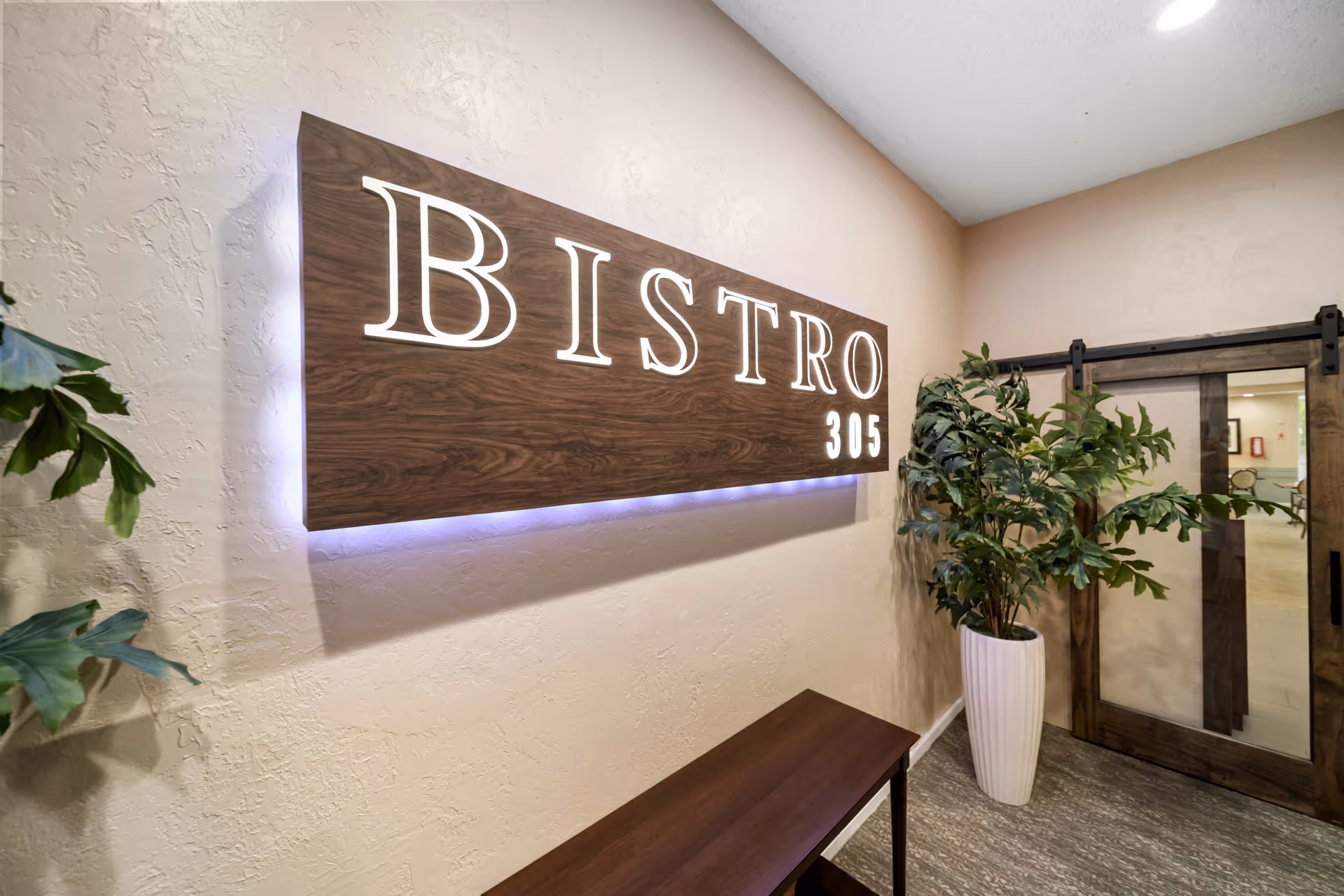 Interior hallway with a wooden sign that reads 'BISTRO 305' illuminated with backlighting, a wooden bench below the sign, a large potted plant to the right, and a sliding barn door with glass panels leading to another room.