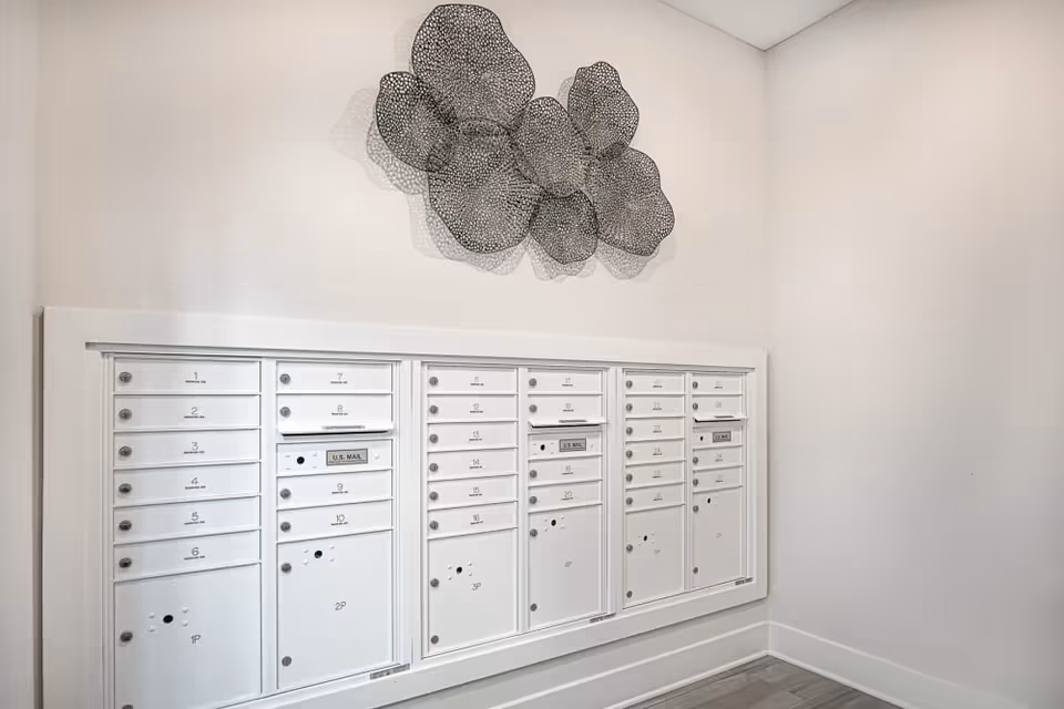 A row of white community mailboxes mounted on a pale wall beneath decorative metal wall art in a hallway.
