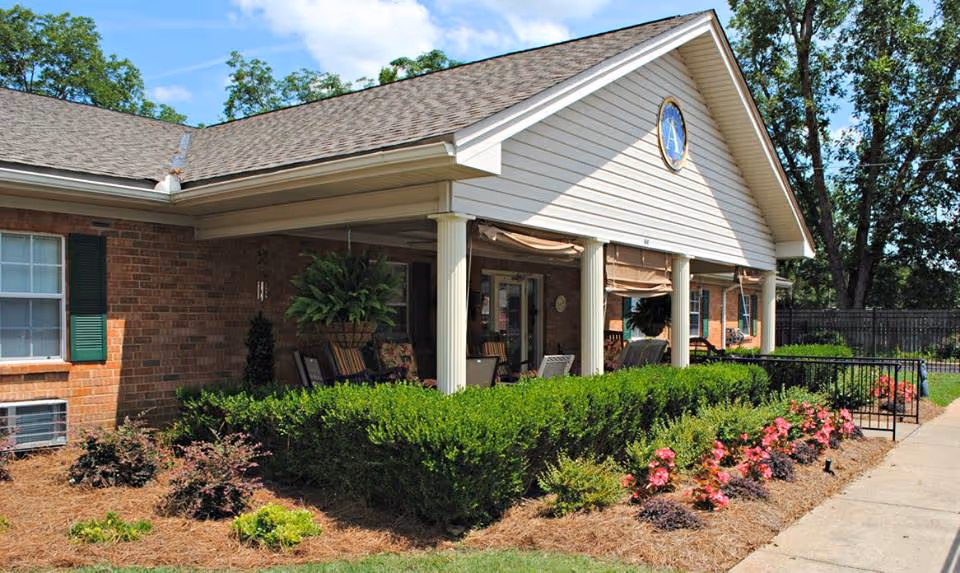 Exterior view of a single-story brick building with a covered porch supported by white columns. The porch has outdoor seating and hanging plants. The building is surrounded by neatly trimmed bushes, colorful flowers, and a paved walkway. Trees and a clear blue sky are visible in the background.