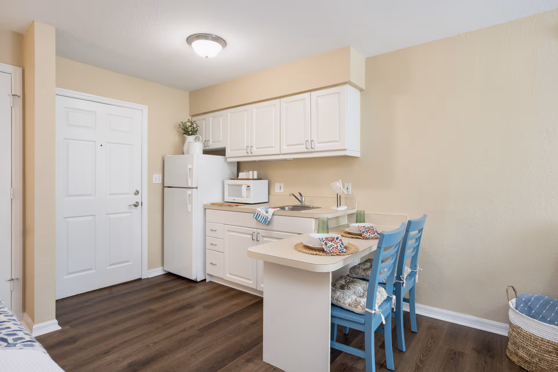 Small kitchen area with white cabinets, a white refrigerator, a microwave on the counter, a sink, and a small dining counter with two blue chairs set with placemats, bowls, and glasses. The floor is dark wood, and the walls are beige. A white door is visible to the left.