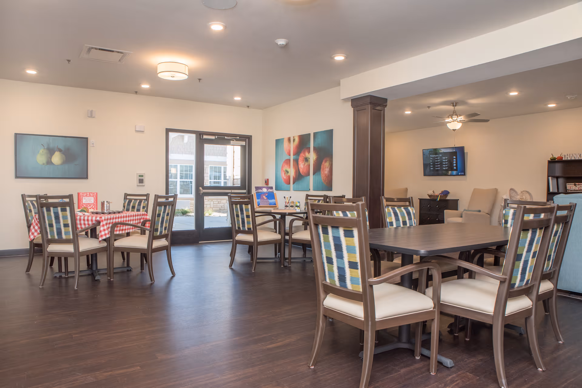 A bright and spacious dining room in a senior living facility with multiple tables and chairs. One table has a red and white checkered tablecloth and a small menu or sign. The walls are decorated with paintings of pears and apples. There is a door with glass panels leading outside, and a seating area with armchairs and a TV mounted on the wall in the background.
