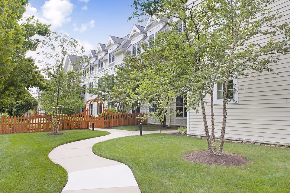 A curved concrete pathway leads through a well-maintained green lawn with several trees and shrubs, alongside a multi-story beige building with white trim and multiple windows under a blue sky with some clouds.
