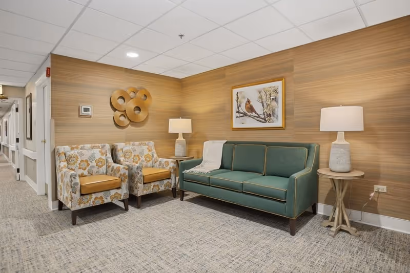 A cozy seating area in a senior living facility hallway featuring a green upholstered sofa with a white throw blanket, two floral-patterned armchairs with tan cushions, two side tables each with a lamp, and wall decorations including a framed bird painting and circular wooden art pieces. The walls have a wood panel design and the floor is carpeted.