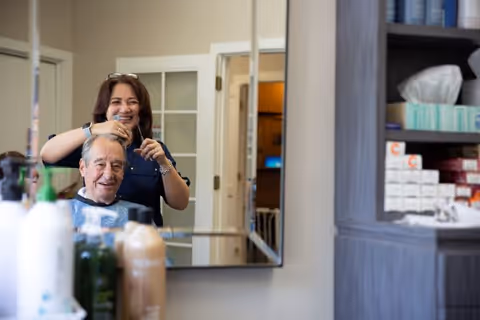 A senior man is getting a haircut from a smiling woman in a salon or barbershop setting. The man is seated and covered with a barber cape, while the woman stands behind him holding scissors. The scene is reflected in a large mirror on the wall. Shelves with various hair care products and supplies are visible to the right.