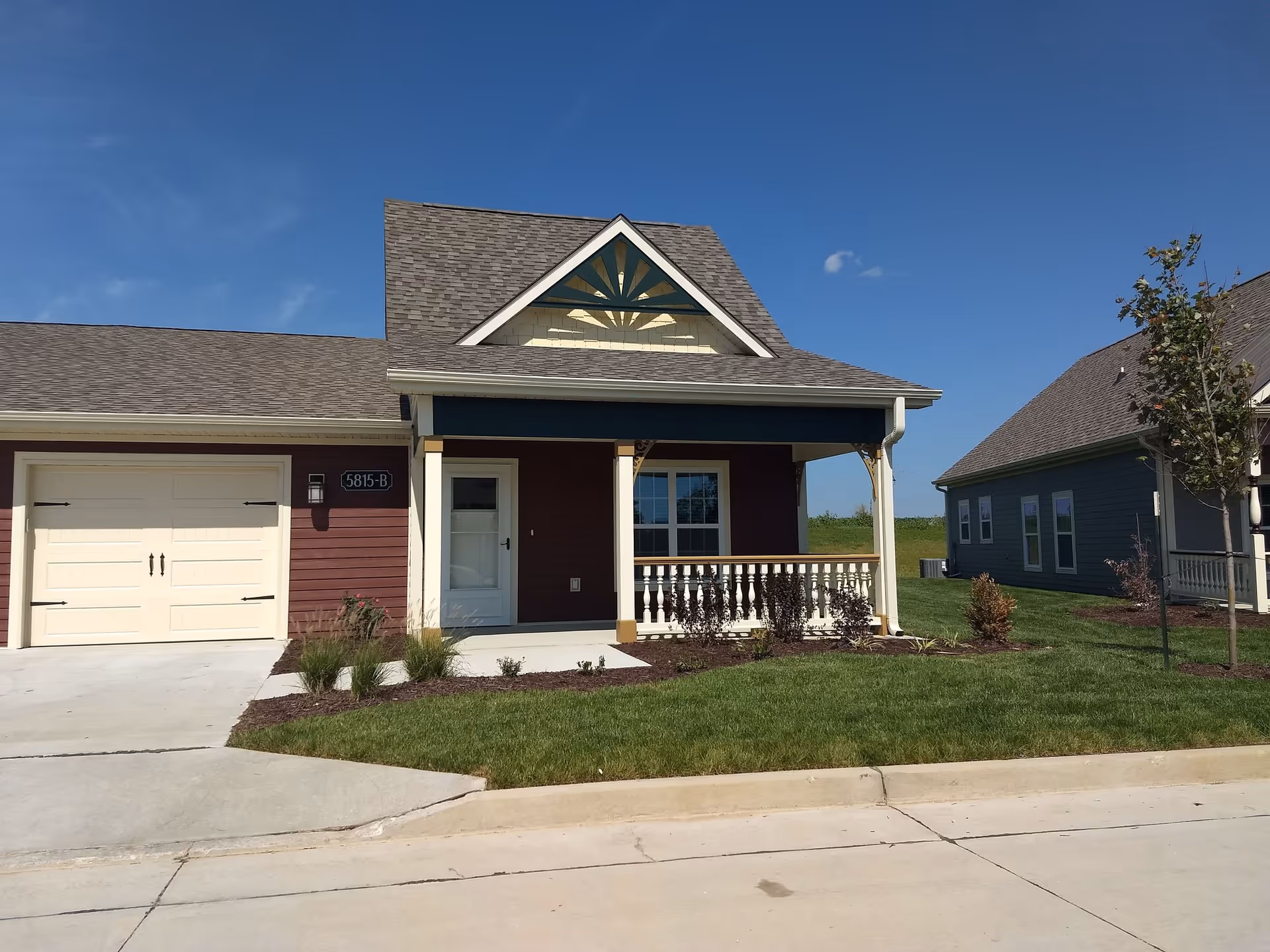 Exterior view of a single-story residential building with a small covered porch, white railing, and a garage door. The building has a gable roof with decorative trim and is surrounded by a well-maintained lawn and small shrubs. Another similar building is visible in the background under a clear blue sky.