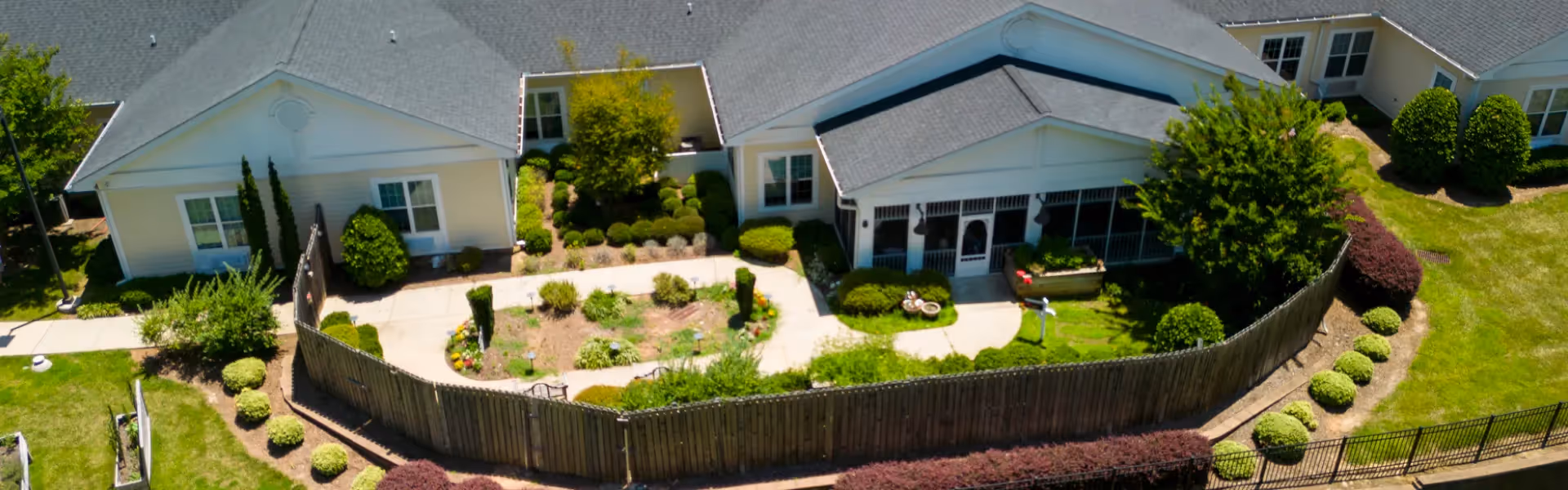 Aerial view of a senior living facility courtyard with a fenced garden area, surrounded by single-story buildings with gray roofs and beige siding. The garden features a walking path, various shrubs, and small trees, with well-maintained landscaping around the buildings.