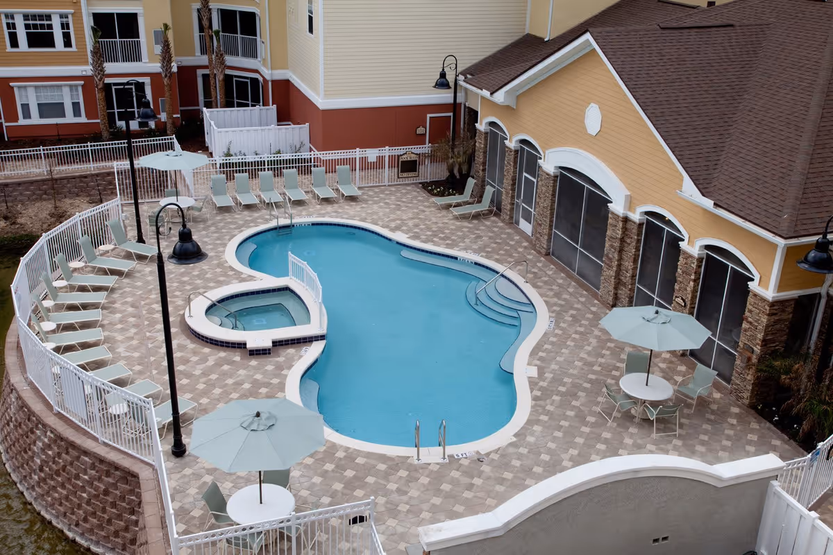 Outdoor swimming pool area at Villa Grande on Saxon with a uniquely shaped pool and a smaller attached hot tub. The pool is surrounded by a tiled deck with multiple lounge chairs and tables with umbrellas. The building adjacent to the pool has large screened windows and a brown shingled roof.