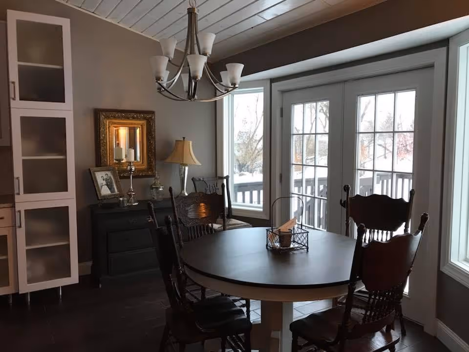 A dining area with a round wooden table surrounded by five dark wooden chairs. The room features large windows and glass doors that lead outside, allowing natural light to fill the space. There is a chandelier hanging from the ceiling above the table. Against the wall, there is a dark sideboard with a lamp, framed photo, decorative candle holder, and a gold-framed mirror. The floor is dark, and the walls are painted a neutral color.