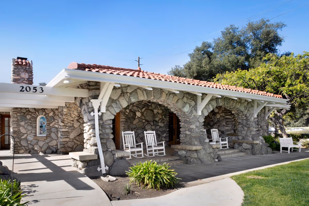 Stone building with a red tile roof and three stone archways on the front porch, each with white rocking chairs. The building number 2053 is visible on a white beam. There is a chimney on the left side and green trees and grass surrounding the building under a clear blue sky.