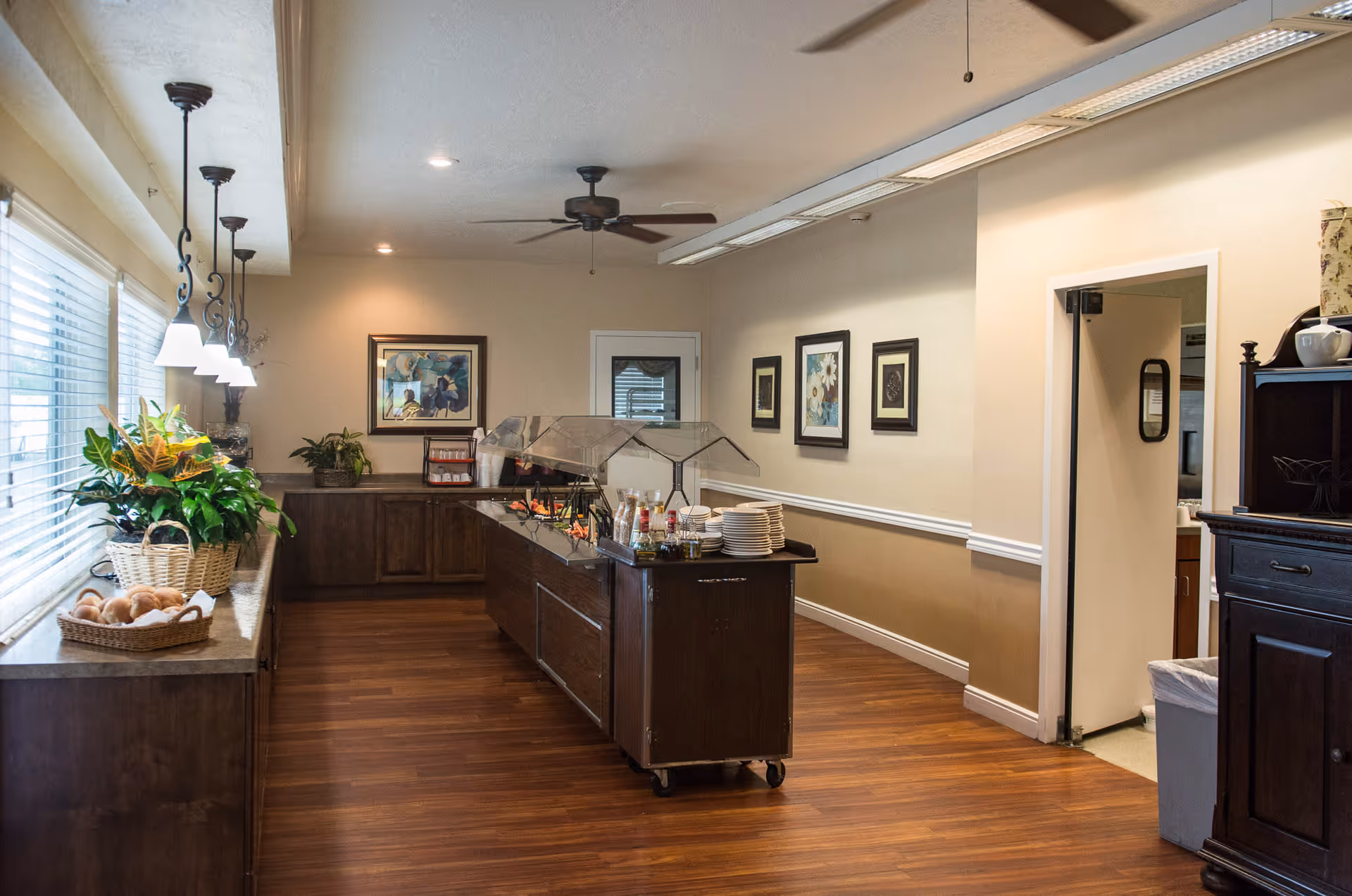 Interior view of a dining area in Highland Cove Retirement featuring a buffet station with plates, condiments, and food under a glass sneeze guard. The room has wooden flooring, beige walls with framed artwork, ceiling fans, and hanging pendant lights over a counter with plants and a basket of bread rolls near large windows.