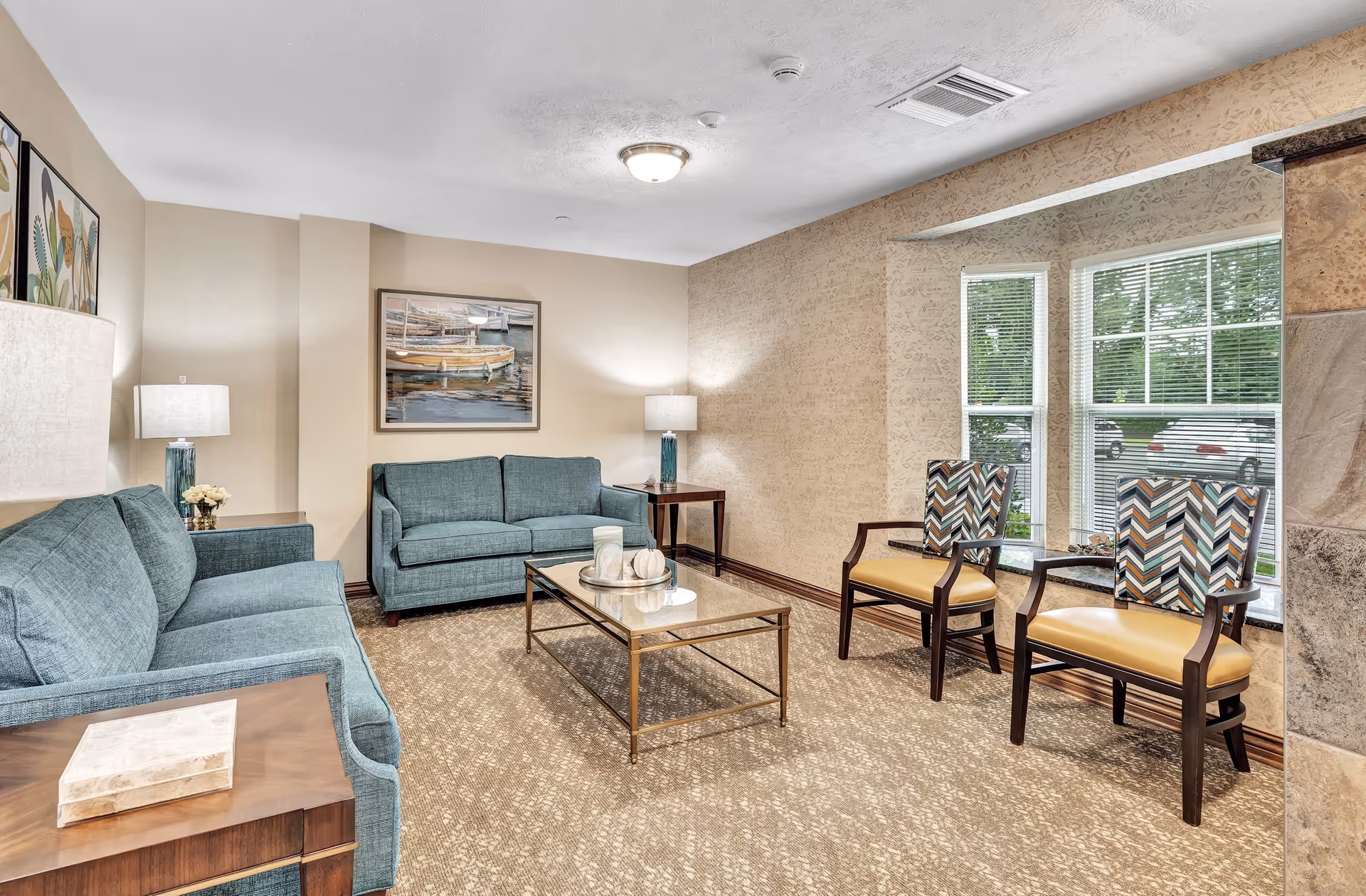 A cozy living room in a retirement community featuring two teal sofas, a glass coffee table with decorative items, two patterned armchairs with yellow seats near a window, beige carpet, and soft lighting from table lamps. The walls are decorated with framed artwork and textured wallpaper.
