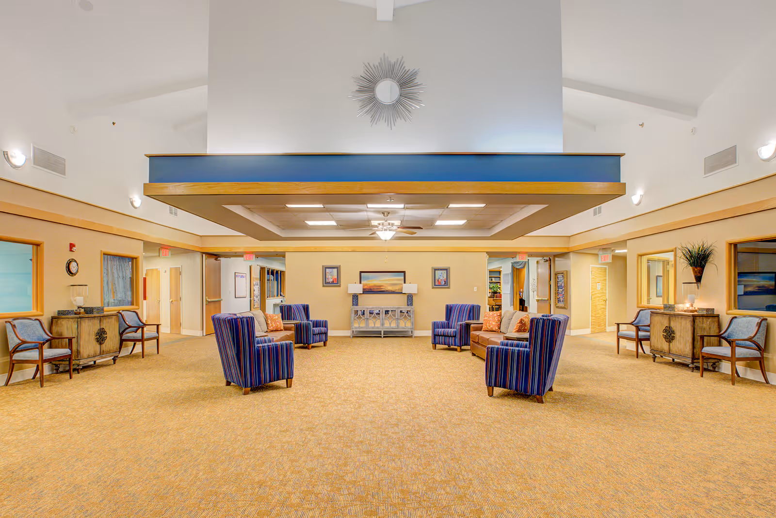 A spacious and well-lit common area with beige carpet and walls. The room features a seating arrangement with four blue striped armchairs and a brown sofa with orange pillows. There are two wooden cabinets with chairs on either side of the room. A decorative sunburst mirror hangs on the white wall above a blue and wood accent structure. The ceiling has recessed lighting and a ceiling fan in the center.