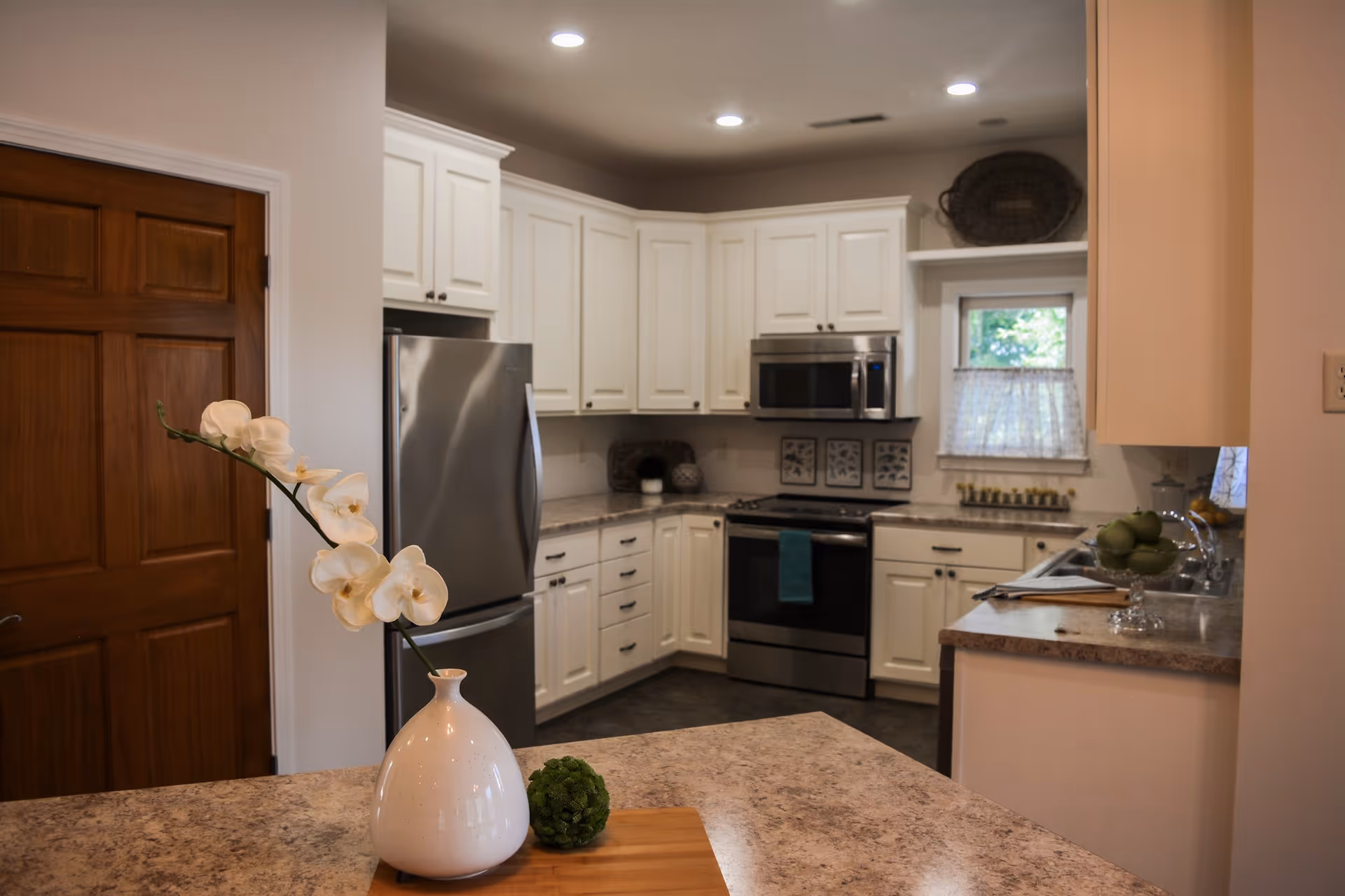 A modern kitchen with white cabinets, stainless steel refrigerator, oven, and microwave. The countertops are a light brown granite, and there is a white vase with white orchids and a small green decorative ball on the counter in the foreground. A wooden door is visible on the left side, and a small window with a lace curtain is above the sink on the right.